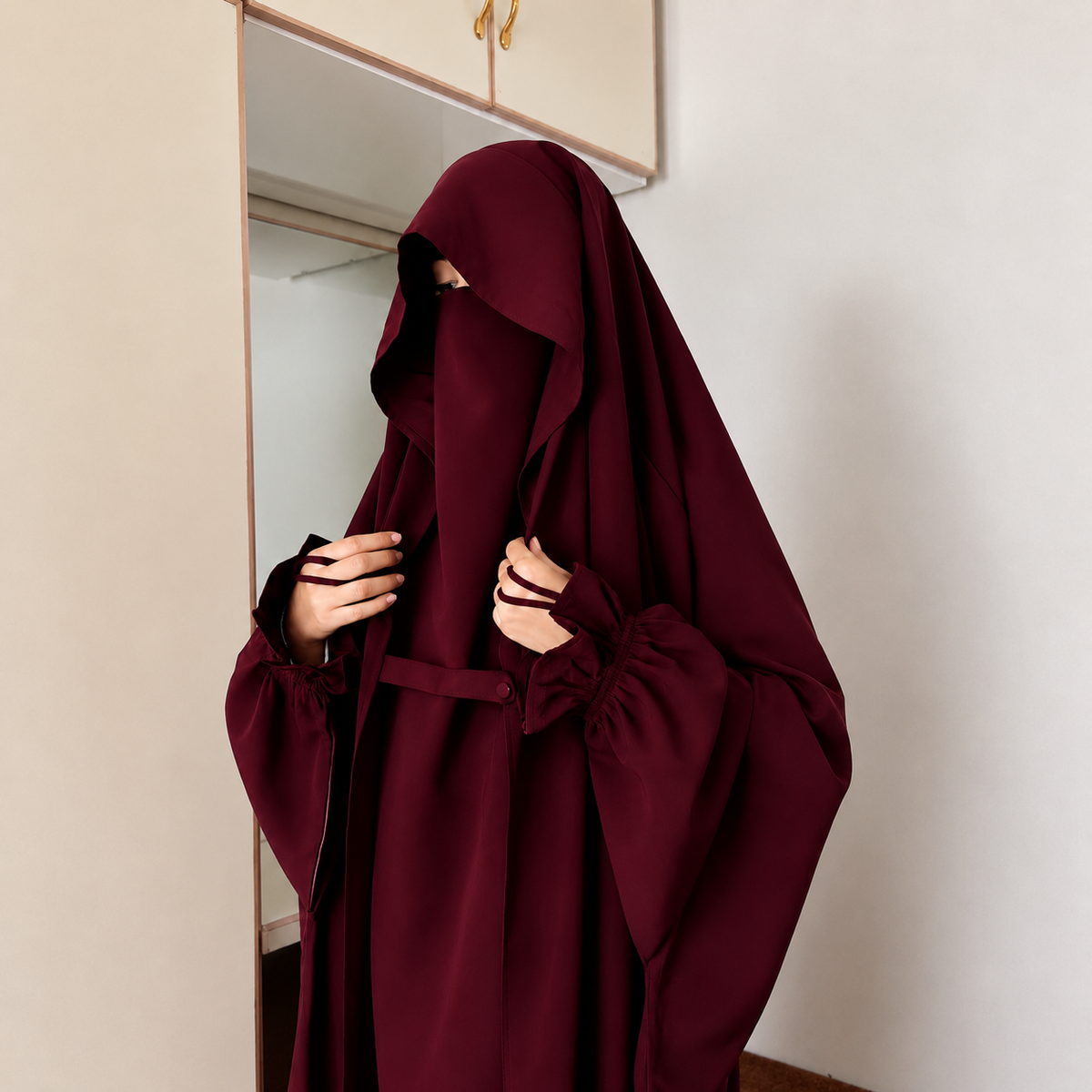 A person wearing a Maroon Saudi Jilbab modest garment with a full face veil and long gathered sleeves, standing indoors near a mirror and wall.