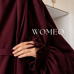 A  person wearing a flowing Saudi Jilbab or abaya with gathered sleeves . A hand is visible holding the fabric, and the brand name 'WOMEO' appears in white text across the center.